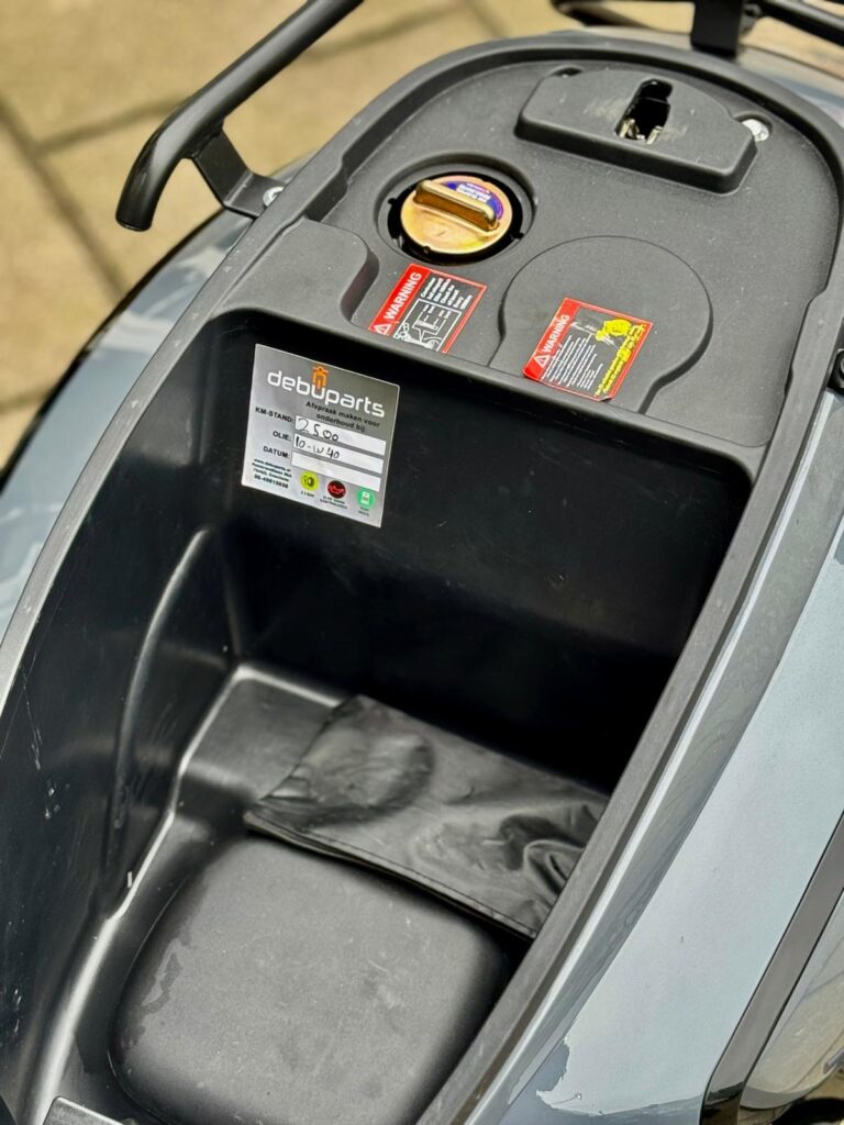 Opbergvak onder het zadel van een scooter met tankdop, waarschuwingstickers en service-label.
