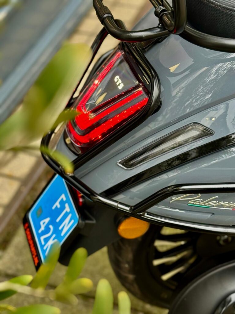 Close-up van de achterkant van een grijze scooter met rood achterlicht, bagagerek en blauwe kentekenplaat.
