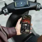 Hand met smartphone die een e-scooter app toont, voor het stuur en display van een scooter.
