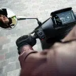 Close-up van een scooterstuur met handschoen, digitaal dashboard en spiegel met berijder.