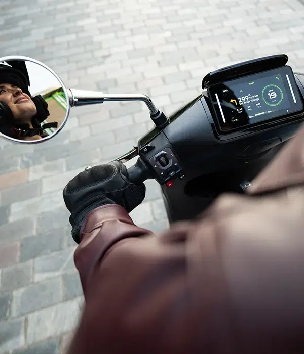 Close-up van een scooterstuur met handschoen, digitaal dashboard en spiegel met berijder.
