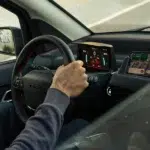 Hand aan het stuur van een elektrische auto met digitaal dashboard en navigatie op een smartphone.