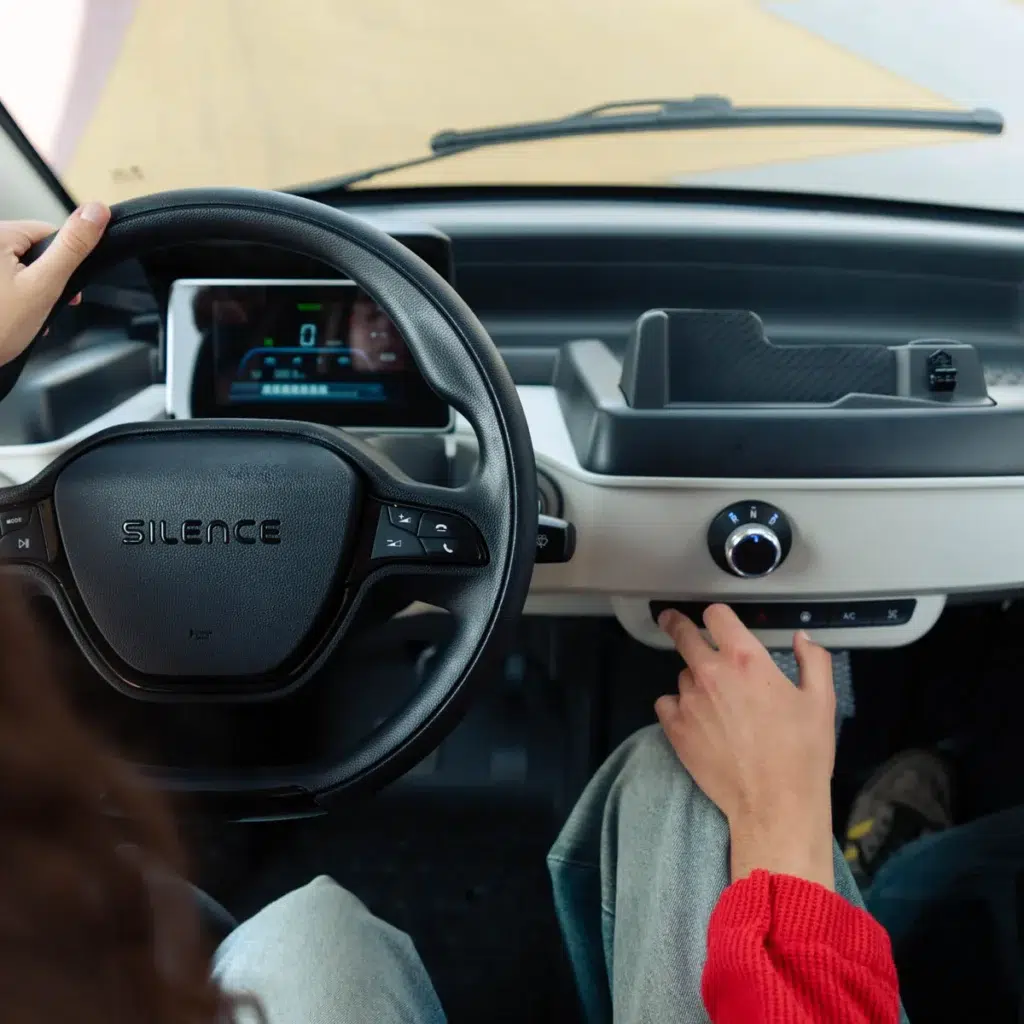 Interieur van een compacte elektrische auto: stuur met Silence-logo, digitaal scherm en hand bij klimaatbediening.