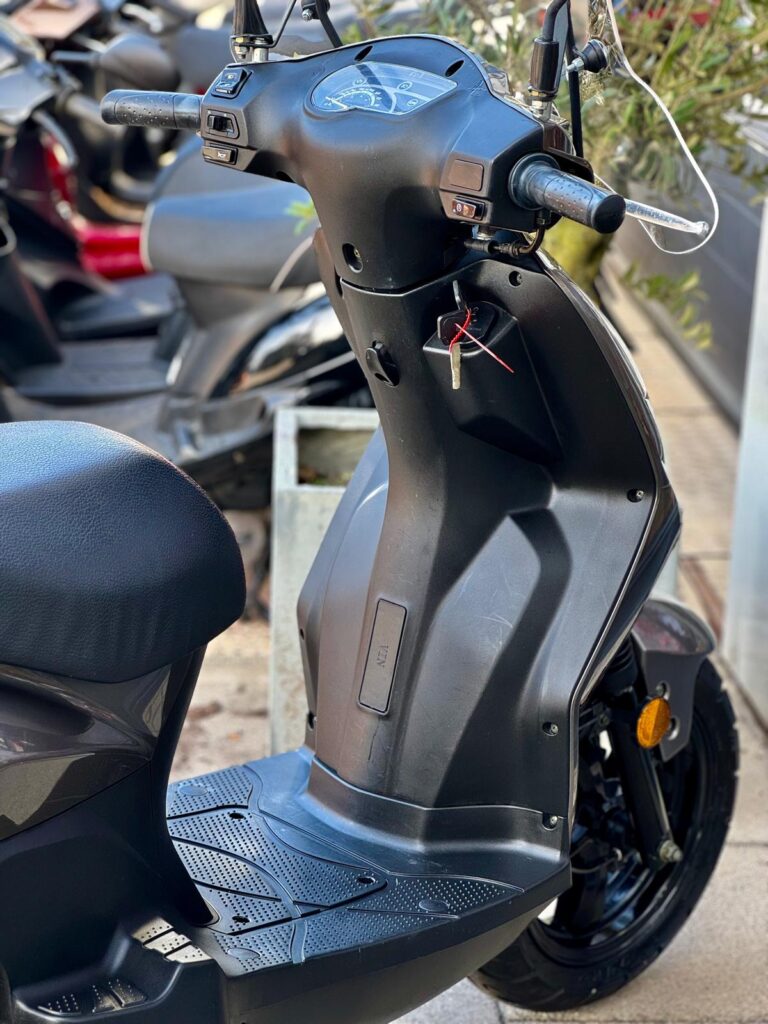 Close-up van het stuur, dashboard en voetplaat van een zwarte scooter met sleutel, geparkeerd buiten.