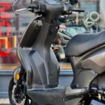 Close-up van een grijze scooter bij een showroom, met stuur, voetplaat en voorwiel zichtbaar.