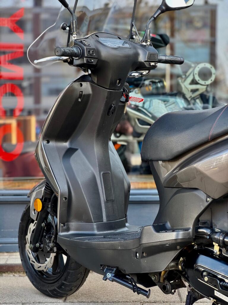 Close-up van een grijze scooter bij een showroom, met stuur, voetplaat en voorwiel zichtbaar.