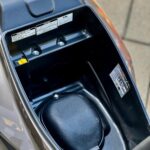 Open opbergruimte onder het zadel van een scooter, met stickers en zwart afdekkapje.