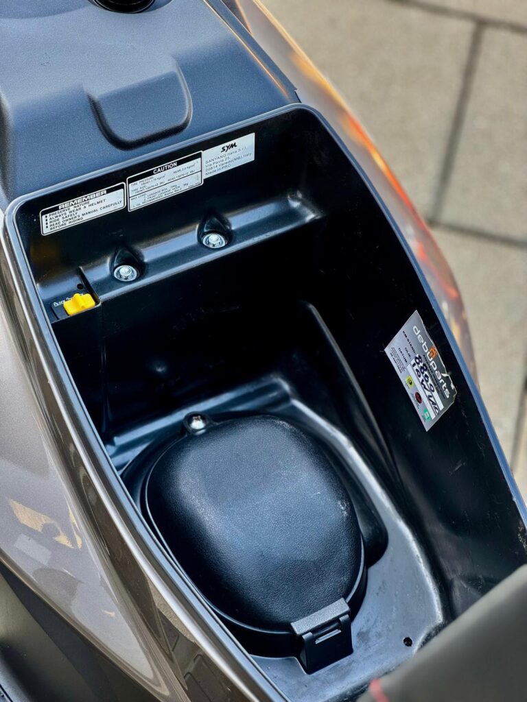 Open opbergruimte onder het zadel van een scooter, met stickers en zwart afdekkapje.