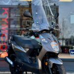 Grijze Kymco-scooter met hoog windscherm, op de standaard voor een winkel met glazen pui.