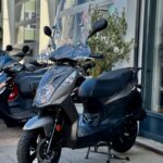 Grijze scooter met hoog windscherm, geparkeerd voor een winkel; andere scooters op de achtergrond.