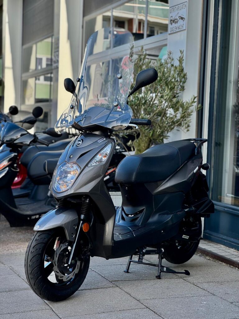 Grijze scooter met hoog windscherm, geparkeerd voor een winkel; andere scooters op de achtergrond.