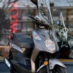 Grijze SYM-scooter met windscherm op de standaard voor een winkelraam.