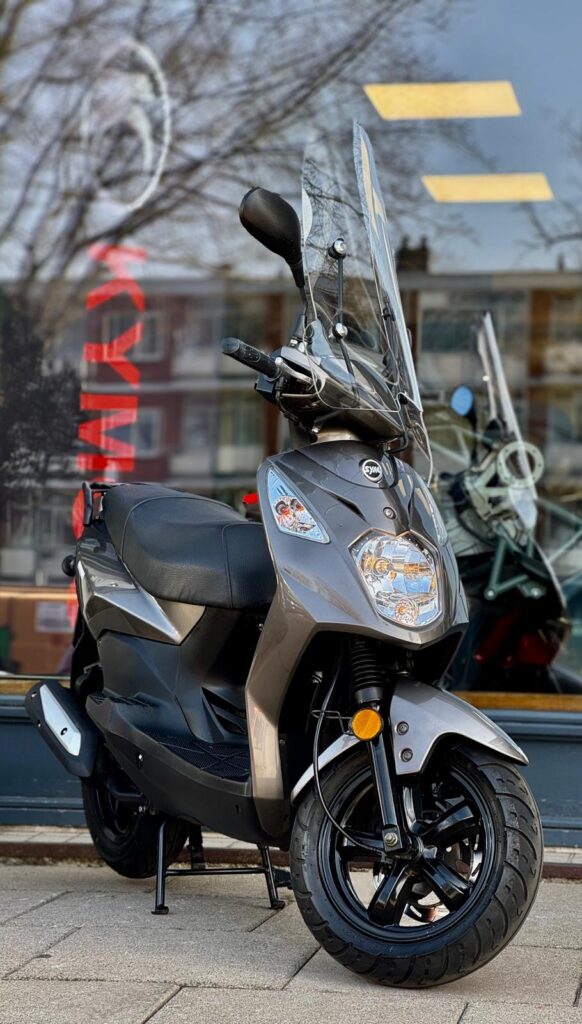 Grijze SYM-scooter met windscherm op de standaard voor een winkelraam.