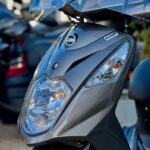 Close-up van de voorzijde van een grijze SYM-scooter met koplamp en ruit, Euro 4-sticker zichtbaar.