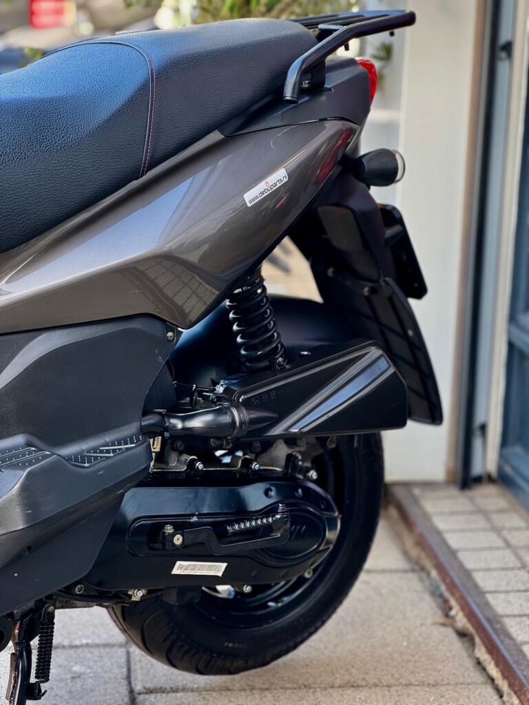 Close-up van de achterzijde van een grijze scooter met zadel, veer en achterwiel op de stoep.