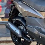 Close-up van de achterzijde van een grijze scooter met zadel, uitlaat en achterwiel op standaard.