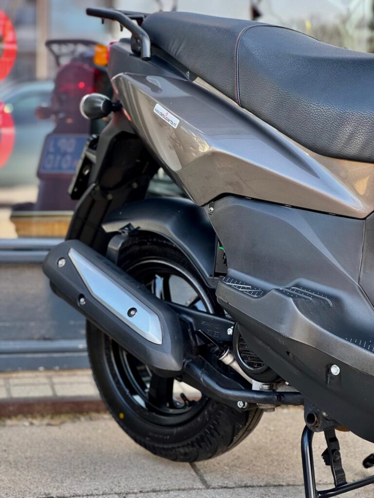 Close-up van de achterzijde van een grijze scooter met zadel, uitlaat en achterwiel op standaard.