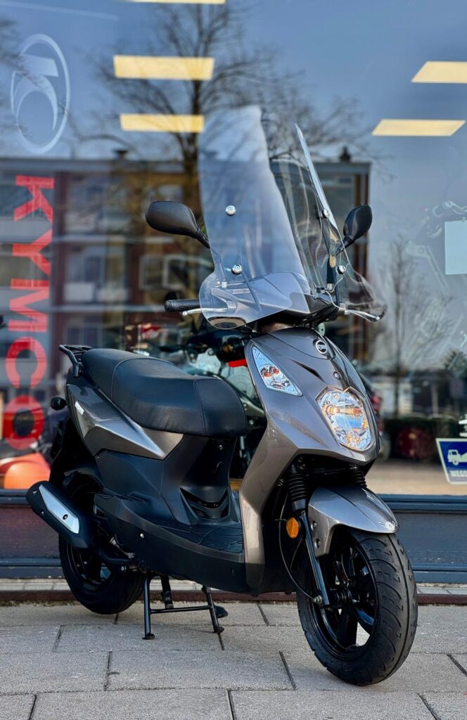 Grijze Kymco-scooter met hoog windscherm, op de standaard voor een winkel met glazen pui.