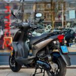 Grijs-zwarte scooter met ruitje geparkeerd voor een showroom met glazen pui.
