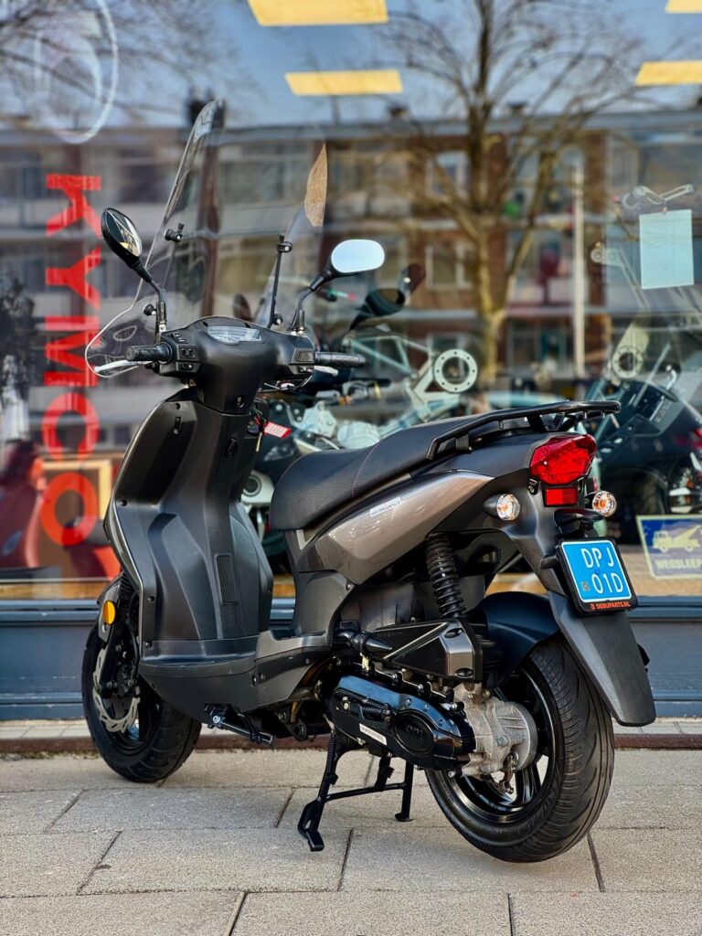 Grijs-zwarte scooter met ruitje geparkeerd voor een showroom met glazen pui.