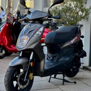 Grijze scooter op de middenstandaard voor een showroom, met rode scooters vervaagd op de achtergrond.