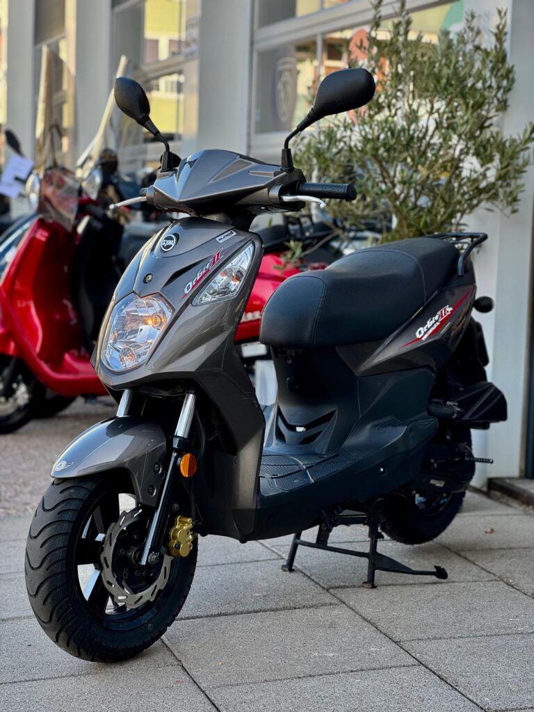Grijze scooter op de middenstandaard voor een showroom, met rode scooters vervaagd op de achtergrond.