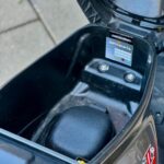 Open zadelbak van een scooter met lege opbergruimte en onderdelensticker.