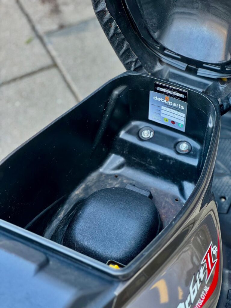Open zadelbak van een scooter met lege opbergruimte en onderdelensticker.