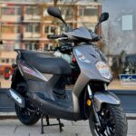 Grijze scooter op standaard voor een winkelraam, met flats weerspiegeld op de achtergrond.