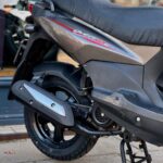 Achterzijde van een grijze scooter op de standaard, met achterwiel en uitlaat, geparkeerd op de stoep.