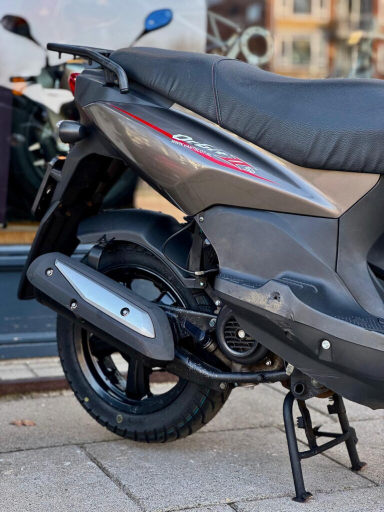 Achterzijde van een grijze scooter op de standaard, met achterwiel en uitlaat, geparkeerd op de stoep.