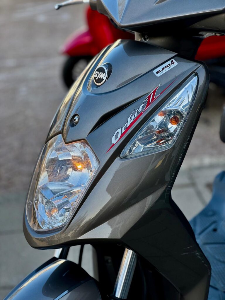 Close-up van de voorkant van een grijze SYM Orbit II scooter met koplampen.