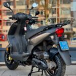 Grijze motorscooter op standaard met blauwe kentekenplaat, geparkeerd voor een winkelraam.