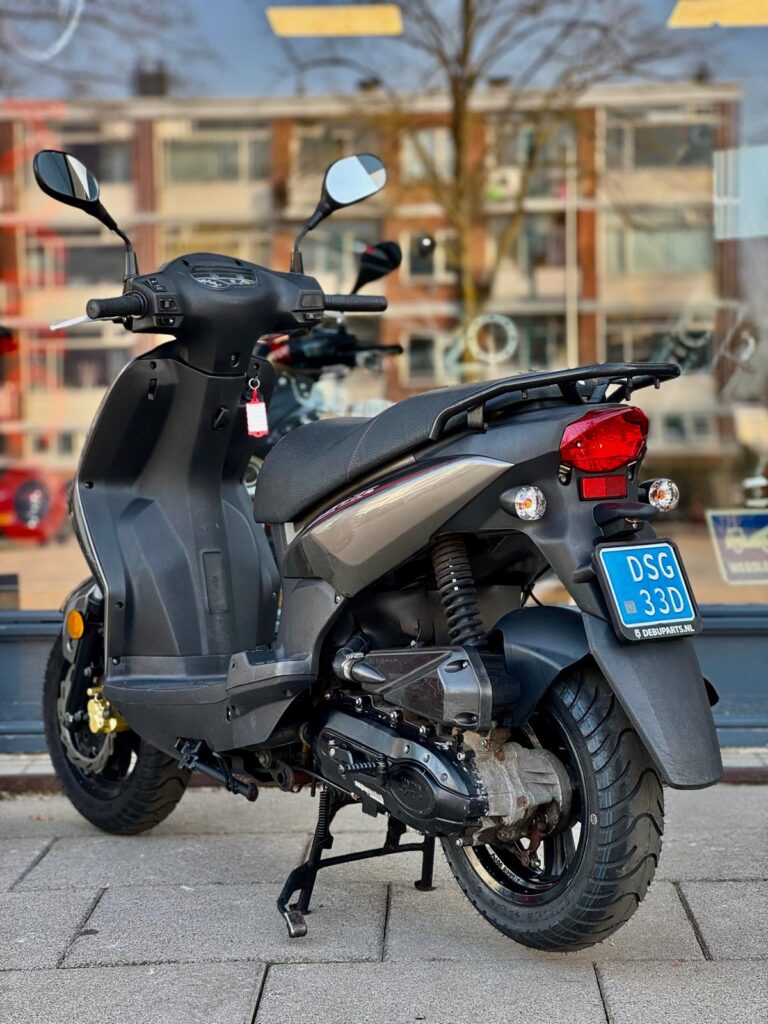 Grijze motorscooter op standaard met blauwe kentekenplaat, geparkeerd voor een winkelraam.