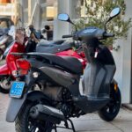 Grijze scooter met blauwe kentekenplaat geparkeerd op de stoep bij een winkel, met andere scooters op de achtergrond.