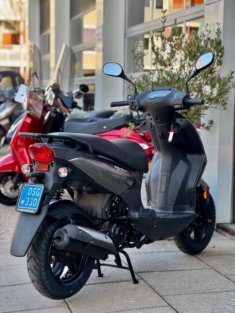 Grijze scooter met blauwe kentekenplaat geparkeerd op de stoep bij een winkel, met andere scooters op de achtergrond.
