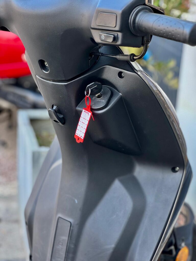 Close-up van het stuur en dashboard van een scooter met sleutel en rood label in het contactslot.