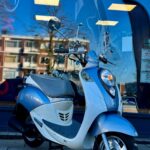 Lichtblauwe retro scooter met windscherm geparkeerd voor een winkelraam met reflectie van flats en blauwe lucht.