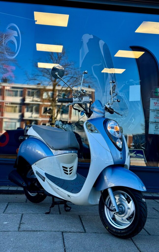 Lichtblauwe retro scooter met windscherm geparkeerd voor een winkelraam met reflectie van flats en blauwe lucht.