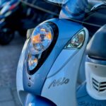 Close-up van de voorkant van een blauwe SYM Mio scooter met brandende koplamp en knipperlicht.