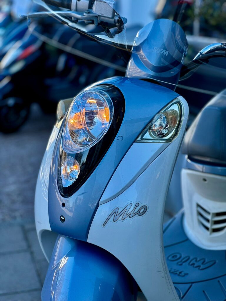 Close-up van de voorkant van een blauwe SYM Mio scooter met brandende koplamp en knipperlicht.