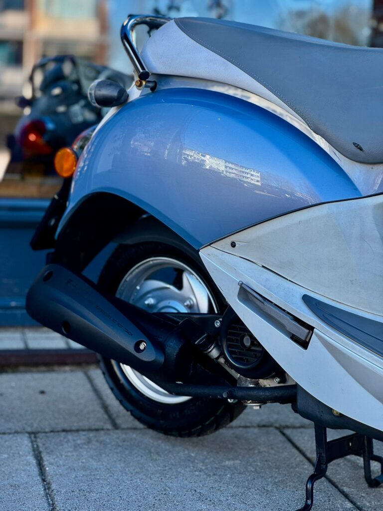 Close-up van de achterkant van een lichtblauwe scooter met grijze zitting en uitlaat op de stoep.