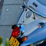Close-up van de achterkant van een blauwe SYM-scooter met rood achterlicht en geel kenteken.