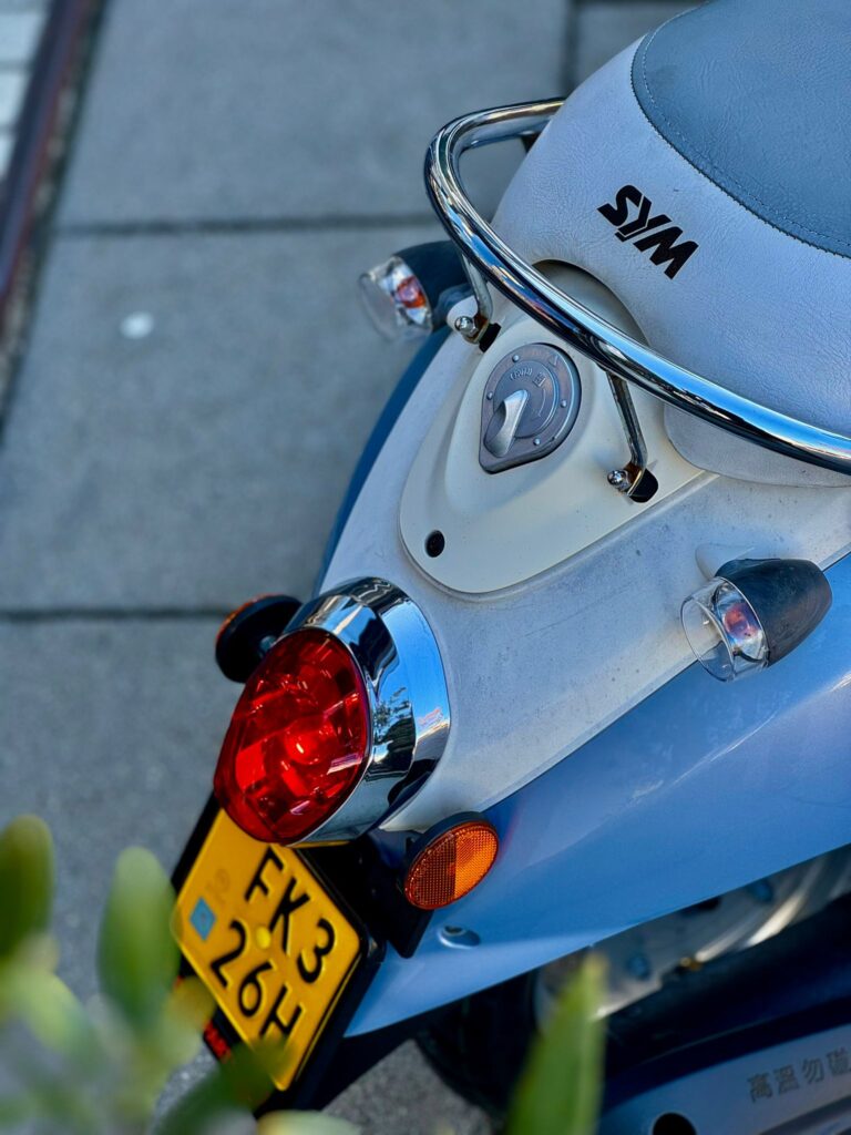 Close-up van de achterkant van een blauwe SYM-scooter met rood achterlicht en geel kenteken.
