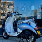 Blauw-witte retro scooter geparkeerd op trottoir voor winkelruit met reflecties.