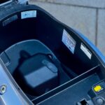 Open zadelbak van een scooter met lege opbergruimte en stickers binnenin.