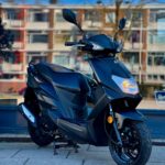 Zwarte scooter geparkeerd op stoep voor winkelraam met flats op de achtergrond.