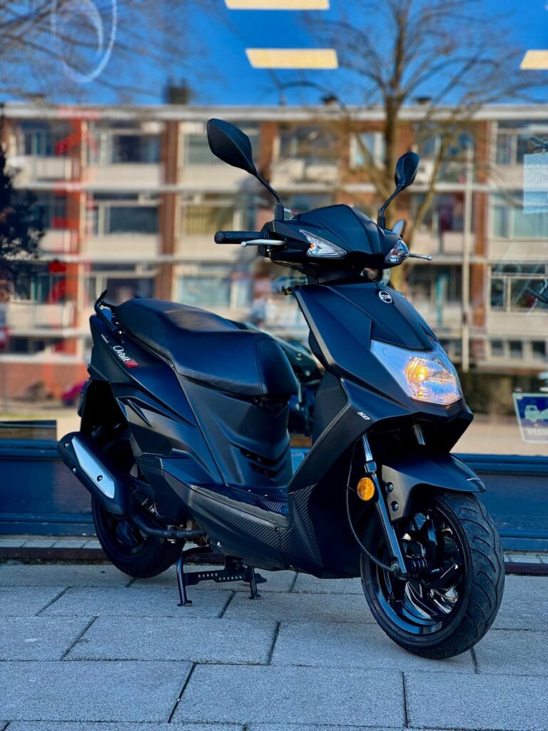 Zwarte scooter geparkeerd op stoep voor winkelraam met flats op de achtergrond.