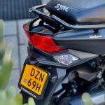 Achterkant van een zwarte SYM-scooter met rood achterlicht en gele Nederlandse kentekenplaat, geparkeerd op straat.