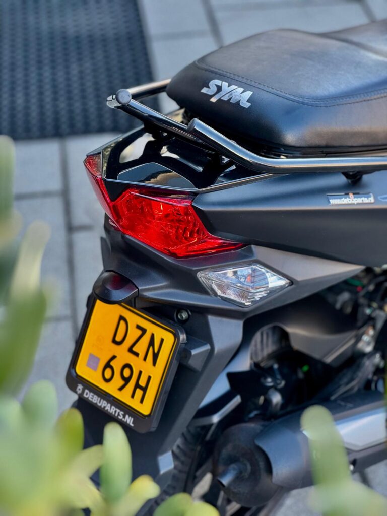 Achterkant van een zwarte SYM-scooter met rood achterlicht en gele Nederlandse kentekenplaat, geparkeerd op straat.